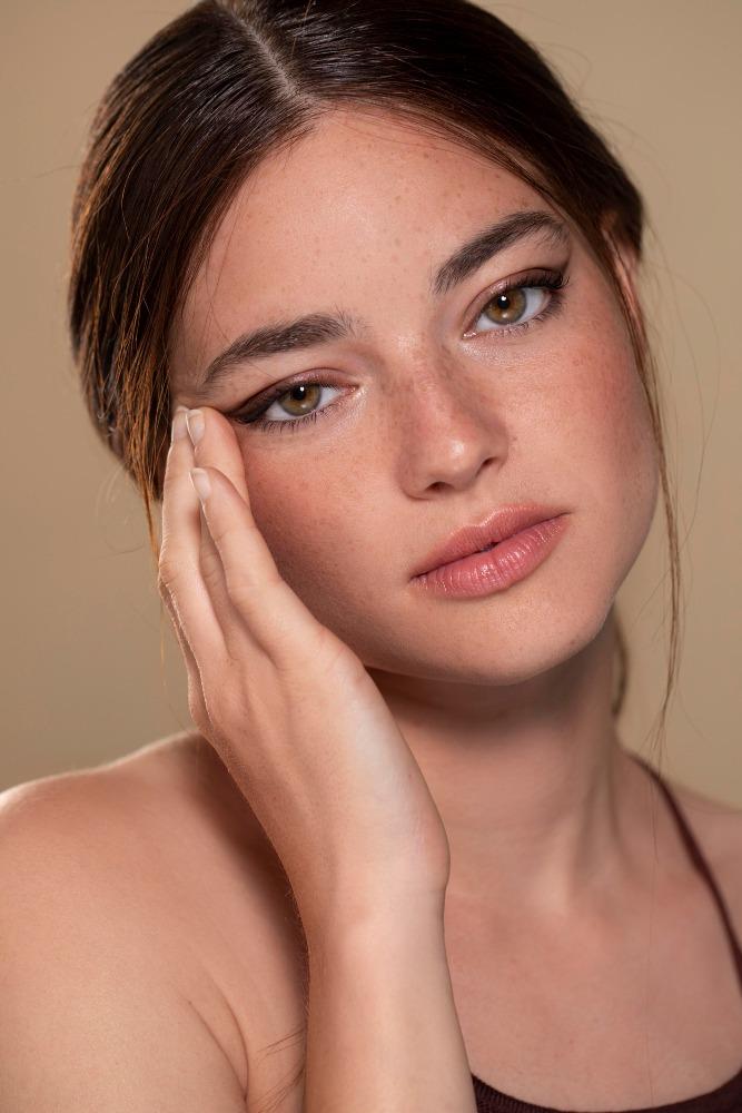 Nahaufnahme einer jungen Frau mit Sommersprossen und dezentem Make-up, die ihre Wange mit der Hand berührt und direkt in die Kamera blickt.