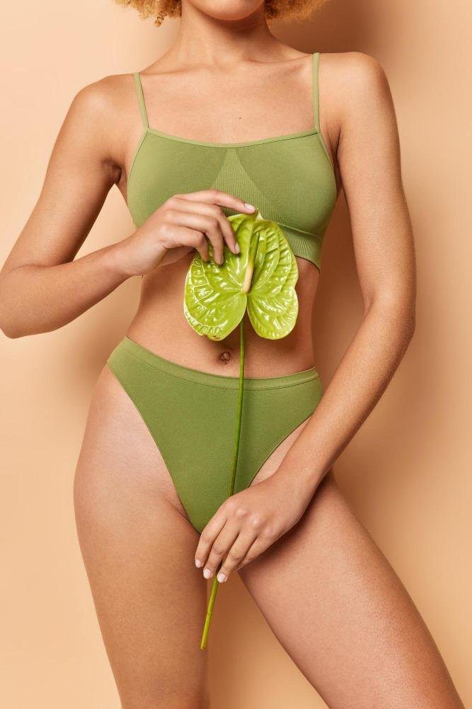 Frau in grüner Unterwäsche hält ein grünes Anthurium-Blatt vor dem Unterbauch, beige Hintergrund.