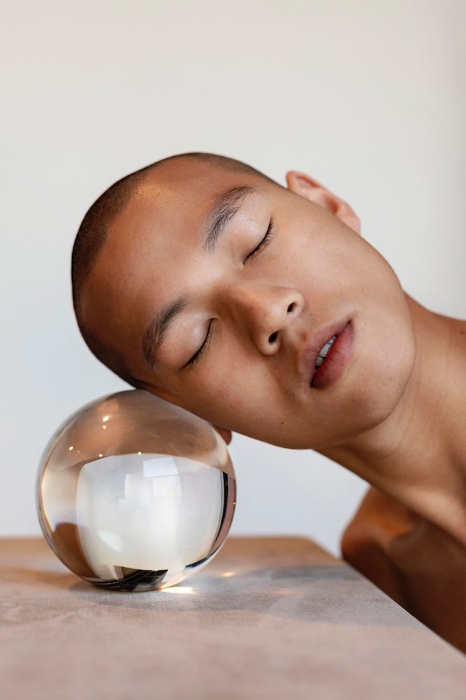 Person with shaved head resting their face gently against a reflective glass sphere on a table, eyes closed in a serene expression.
