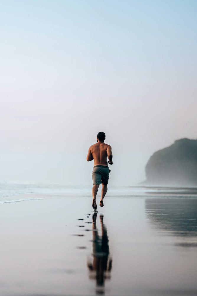 Man jogging shirtless on a beach during sunrise with reflection on wet sand.