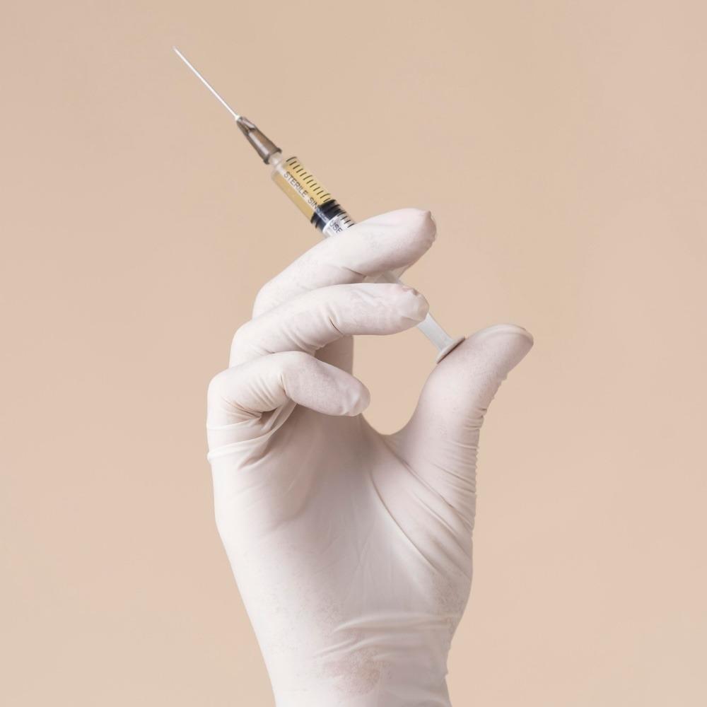 Gloved hand holding a syringe filled with yellowish liquid against a neutral beige background.
