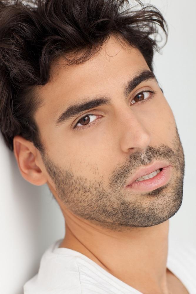 Porträt eines jungen Mannes mit dunklen Haaren und Bart, der in die Kamera blickt, während er in einem weißen Shirt vor neutralem Hintergrund posiert.