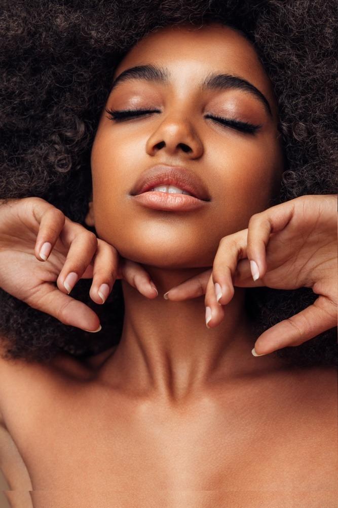 Nahaufnahme einer Frau mit geschlossenen Augen und natürlichem Make-up, die ihr Gesicht entspannt mit beiden Händen sanft berührt.