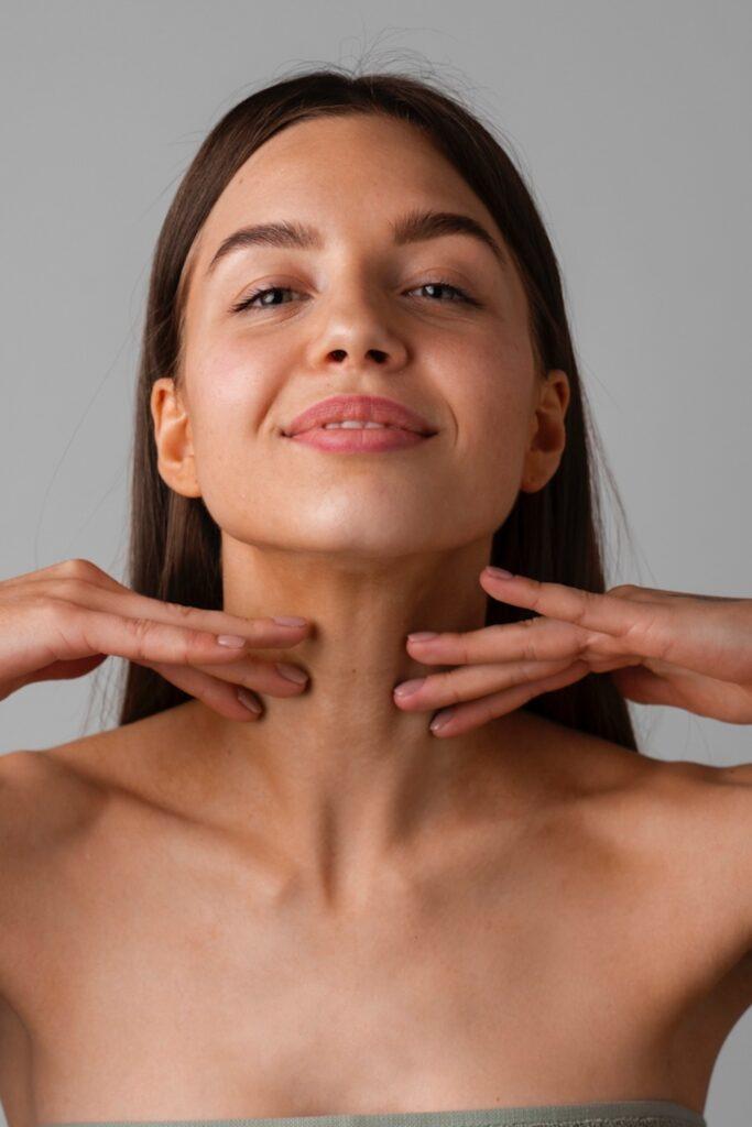 Frau mit leichtem Lächeln legt beide Hände unter das Kinn und betont den Hals vor grauem Hintergrund.