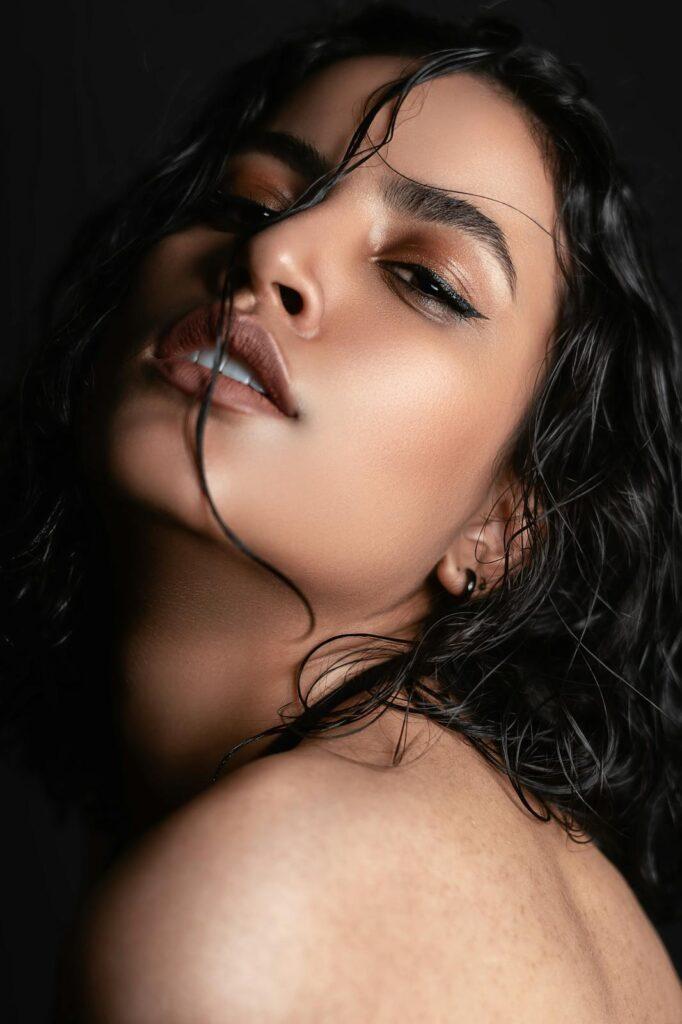 Close-up portrait of a person with wet, wavy hair partially covering their face, wearing bold eyeliner and nude lipstick, gazing intensely at the camera against a dark background.