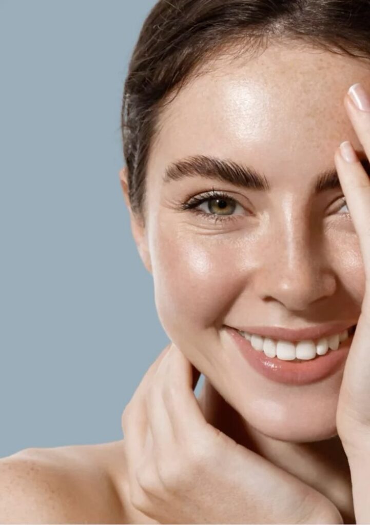 Nahaufnahme einer lächelnden Frau mit taufrischer Haut, die ihr Gesicht mit einer Hand berührt, vor blau-grauem Hintergrund.