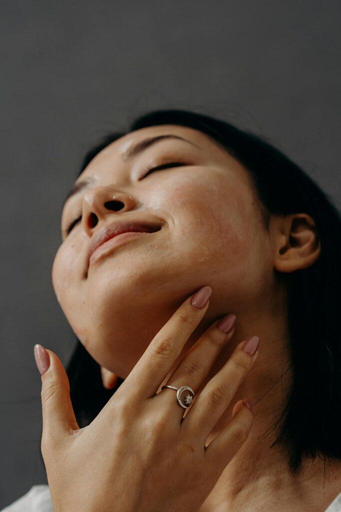 Close-up of a woman with eyes closed, tilting her head back and touching her chin with her hand, showing natural skin texture and a silver ring on her finger.
