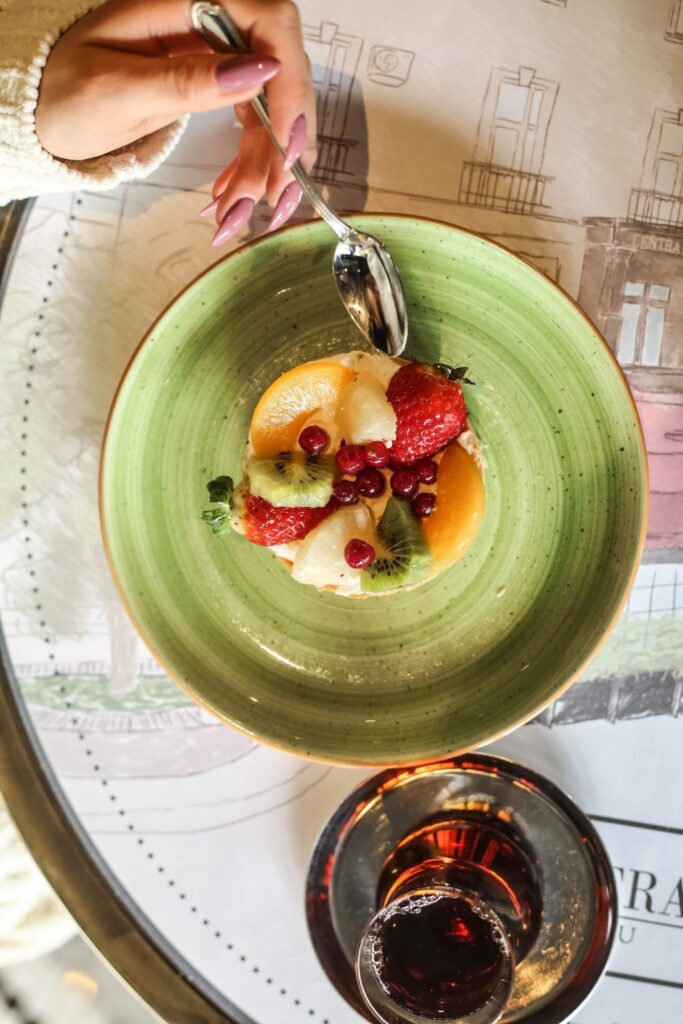 Draufsicht: Hand mit Löffel über einem grünen Teller mit Obsttörtchen (Erdbeeren, Kiwi, Pfirsich, Johannisbeeren); unten ein Glas Tee.